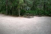 View of a vacant campsite with a fire ring/grill, picnic table, electric pedestal, and a water spigot. The main part of the site is sand. There is grass in the foreground and the back of the site is shaded and lined with trees.