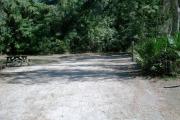 View of site number 11 from the circle drive looking toward the rear of the campsite. You can see the wooden picnic table and fire ring/grill along with the electrical can water hookups to the right. The site is bordered with small and medium sized oak and pine trees with larger trees further back.