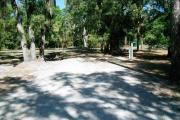 This picture depicts a view from the circle drive looking toward the back of the campsite. You can see the wooden picnic table and fire ring/grill to the left and the electrical and water connections on the right. The campsite is mostly open and has partial shade.
