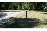 This picture shows the numbered post for campsite 18 which is a 69 foot pull through site. There is a wooden table and a ground fire ring/grill in the background. In the left corner is the electric and water hook ups. It is an open site with grassy areas, saw palmettos and a small magnolia to the right.