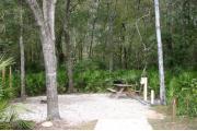 Photo shows an empty gravel pad with trees and vegetation surrounding the site. Site is a short distance from the playground, pavilion, parking, and the bathhouse.