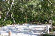 Photo shows an empty gravel pad with trees and vegetation surrounding the site. Site is a short distance from the playground, pavilion, parking, and the bathhouse.