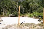 Photo shows an empty gravel pad with trees and vegetation surrounding the site. Site is a short distance from the playground, pavilion, parking, and the bathhouse.