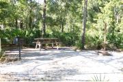Photo shows an empty gravel pad with trees and vegetation surrounding the site. Site is a short distance from the playground, pavilion, parking, and the bathhouse.