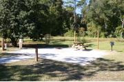 Photo shows an empty gravel pad with a couple small trees. There is only grass around the rest of the site. Site is a short distance from the playground, pavilion, parking, and the bathhouse.