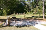 Photo shows an empty gravel pad with a couple small trees. There is only grass around the rest of the site. Site is a short distance from the playground, pavilion, parking, and the bathhouse.