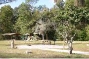 Photo shows an empty gravel pad with playground and pavilion visible through trees a couple small trees. There is only grass around the rest of the site. Site is a short distance from parking and the bathhouse.