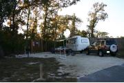 Photo shows a camperâs tow-behind trailer, vehicle, and camping gear on the gravel pad. Photo shows some trees in the background and offers morning shade. This site is in front of wooden privacy fence.