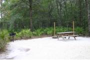This photo shows a campers fire and gear on a gravel pad with trees and vegetation surrounding the site.