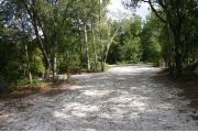 Photo shows a pull through gravel site with trees on both sides giving shade. The photo shows a camperâs RV with the awning out, 2 vehicles, kayaks, and gear.