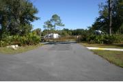 This photo shows an empty paved ADA accessible site with grass and some trees and vegetation surrounding the site. Another RV is visible. This site is a short distance to the bathhouse.