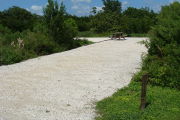Photo: 004, View of gravel campsite with picnic table and grill, Bushes in background.