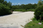 Photo: 005, View of gravel campsite with picnic table and grill, bushes in background.