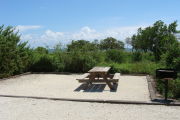 Photo: 008, View of gravel campsite with picnic table and grill, bushes in background.