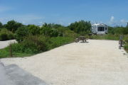 Photo: 014, View of gravel campsite with picnic table and grill, RV and bushes in background.