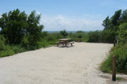 Photo: 019, View of gravel campsite with picnic table and grill, Atlantic Ocean and bushes in background.