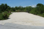 Photo: 020, View of gravel campsite with picnic table and grill, RV and bushes in background.