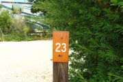 Photo: 023, View of site number post white on brown, bushes and RV visible in the background.