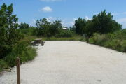 Photo: 028, View of gravel campsite with picnic table and grill. Bushes and trees in background.