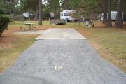 A view of RV site nineteen at Topsail Hill Preserve State Park looking towards the back of the site.  A picnic table is on a concrete pad to the left of the site and utility hook up boxes are to the right of the site.  A small wooden bridge and other RV sites are visible in the background.