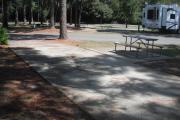 A view of RV site twenty-one at Topsail Hill Preserve State Park looking across the site.  A picnic table is on a concrete pad next to the site.  The site is partially shaded by pine trees.  Other RV sites are visible in the background.