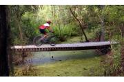 Off-road bike rider speeding across wooden bridge spanning vegetation infested creek.