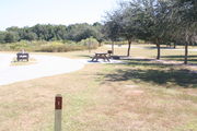 View of campsite #1 from site entry â This is a pull-through campsite, with an asphalt pad for camping units.  On the left of the asphalt pad is the electric hookup and water spigot, surrounded by a protective wooden enclosure.  On the right is a 12â x 20â concrete pad with a picnic table.  Near the concrete pad are a circular ground grill and a pedestal grill.