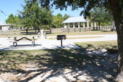 At the back of campsite #1, looking towards campground road - Ground is scattered with green grass and grey soil.  Partial shade is provided by a small oak tree.