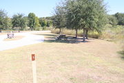 View of campsite #3 from site entry â This is a pull-through campsite, with an asphalt pad for camping units.  On the left of the asphalt pad is the electric hookup and water spigot, surrounded by a protective wooden enclosure.  On the right are a picnic table and a circular ground grill.