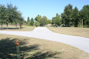 View of campsite #4 from site entry â This is a back-in campsite, with an asphalt pad for camping units.  On the right of the asphalt pad is the electric hookup and water spigot, surrounded by a protective wooden enclosure and a picnic table.  On the left is a circular ground grill.  Partial view of Lake Alafia and its tree lined shore.