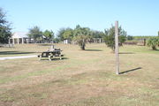Looking at campsite #4 from lake side â Ground cover is green and brown grass, with a view of a pavilion.  A 6â x 6â post marks an area for campers to tie their horses.