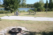 From the side of campsite #4, looking towards Lake Alafia - Ground is scattered with green and brown grass, partial shade is provided by a small oak tree.  Partial view of Lake Alafia and its tree lined shore.