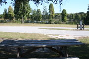 At the back of campsite #5, looking towards campground road - Ground is scattered with green grass and grey soil.  Partial shade is provided by small oak trees.  Partial view of Lake Alafia and its tree lined shore.  Two 6â x 6â posts mark the area for campers to tie their horses.