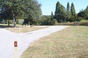 View of campsite #6 from site entry â This is a back-in campsite, with an asphalt pad for camping units.  On the right of the asphalt pad is the electric hookup and water spigot, surrounded by a protective wooden enclosure.  On the left are a circular ground grill and a picnic table, surrounded by small oak trees.  A partial view of Lake Alafia and its tree lined shore is visible.