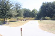 View of campsite #8 from site entry â This is a back-in campsite, with an asphalt pad for camping units.  On the right of the asphalt pad is the electric hookup and water spigot, surrounded by a protective wooden enclosure.  On the left are a circular ground grill and a picnic table.  Partial shade is provided by small oak trees to the left of asphalt pad.