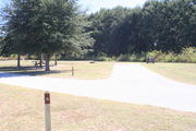 View of campsite #10 from site entry â This is a back-in campsite, with an asphalt pad for camping units.  On the right of the asphalt pad is the electric hookup and water spigot, surrounded by a protective wooden enclosure.  On the left are a circular ground grill and a picnic table.  Partial shade is provided by small oak trees to the left of asphalt pad.
