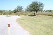 View of campsite #14 from site entry â This is a pull-through campsite, with an asphalt pad for camping units.  On the left of the asphalt pad is the electric hookup and water spigot, surrounded by a protective wooden enclosure.  On the right are a picnic table and a circular ground grill.