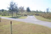 View of campsite #15 from site entry â This is a back-in campsite, with an asphalt pad for camping units.  On the right of the asphalt pad is the electric hookup and water spigot, surrounded by a protective wooden enclosure.  On the left are a circular ground grill and a picnic table.  Brown and green foliage lines the back edge the campsite.