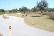 View of campsite #18 from site entry â This is a pull-through campsite, with an asphalt pad for camping units.  On the left of the asphalt pad is the electric hookup and water spigot, surrounded by a protective wooden enclosure.  On the right are a picnic table and a circular ground grill.