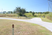 View of campsite #19 from site entry â This is a back-in campsite, with an asphalt pad for camping units.  On the right of the asphalt pad is the electric hookup and water spigot, surrounded by a protective wooden enclosure.  On the left are a circular ground grill and a picnic table.  Green foliage lines the back edge the campsite.