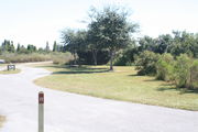 View of campsite #22 from site entry â This is a pull-through campsite, with an asphalt pad for camping units.  On the left of the asphalt pad is the electric hookup and water spigot, surrounded by a protective wooden enclosure.  On the right are a picnic table and a circular ground grill.