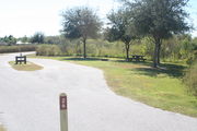 View of campsite #26 from site entry â This is a pull-through campsite, with an asphalt pad for camping units.  On the left of the asphalt pad is the electric hookup and water spigot, surrounded by a protective wooden enclosure.  On the right are a picnic table and a circular ground grill.