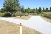 View of campsite #27 from site entry â This is a back-in campsite, with an asphalt pad for camping units.  On the right of the asphalt pad is the electric hookup and water spigot, surrounded by a protective wooden enclosure.  On the left are a circular ground grill and a picnic table, surrounded by small oak trees.  The tree lined shore of Lake Alafia is visible.