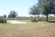 At the back of campsite #27, looking towards campground road - Ground cover is green and brown grass, with patches of grey soil.  On the far right of the photo are a picnic table and circular ground grill.