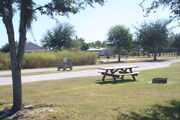 At the back of campsite #28, looking towards campground road - Ground cover is green and brown grass.  Partial shade is provided by small oak trees.  Circular ground grill, picnic table, electric hookup and water spigot are visible in the photo from right to left.