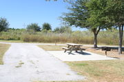 At the back of campsite #29 looking towards road â Ground cover is green and brown grass.  Right side of photo is a picnic table and pedestal grill.