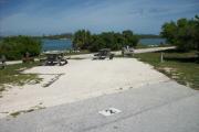 The back has a view of the inlet. The front is open to the street. There is a gravel drive in the center of the site. There is a fire ring next to a picnic table on the left.