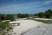 The back has a view of the inlet. The front is open to the street. There is a gravel drive in the center of the site. The left side has a small tree leaning next to a picnic table. The right side has an open grassy area.