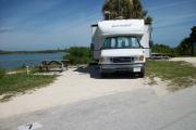 The back has a view of the inlet. The front is open to the street. There is a gravel drive in the center and a grassy area on the right and left sides. A white RV is in the driveway. A picnic table with a fire ring behind it is on the left.