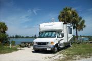 The back has a view of the inlet. The front is open to the street. There is a gravel drive in the center of the site.  There is a picnic table on the left and a white RV with palms behind it in the driveway.